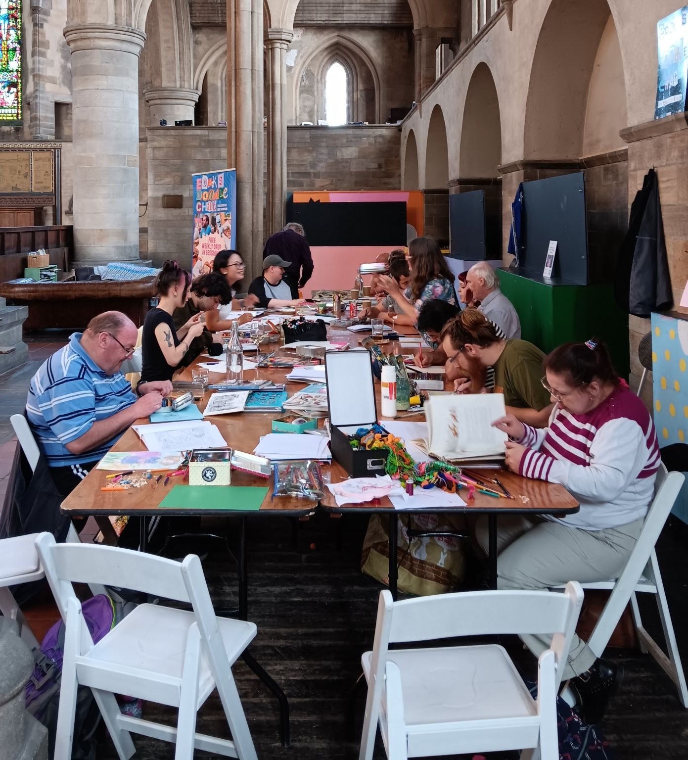 People sat around a big table with lots of colour paper, colours. They are all drawing;